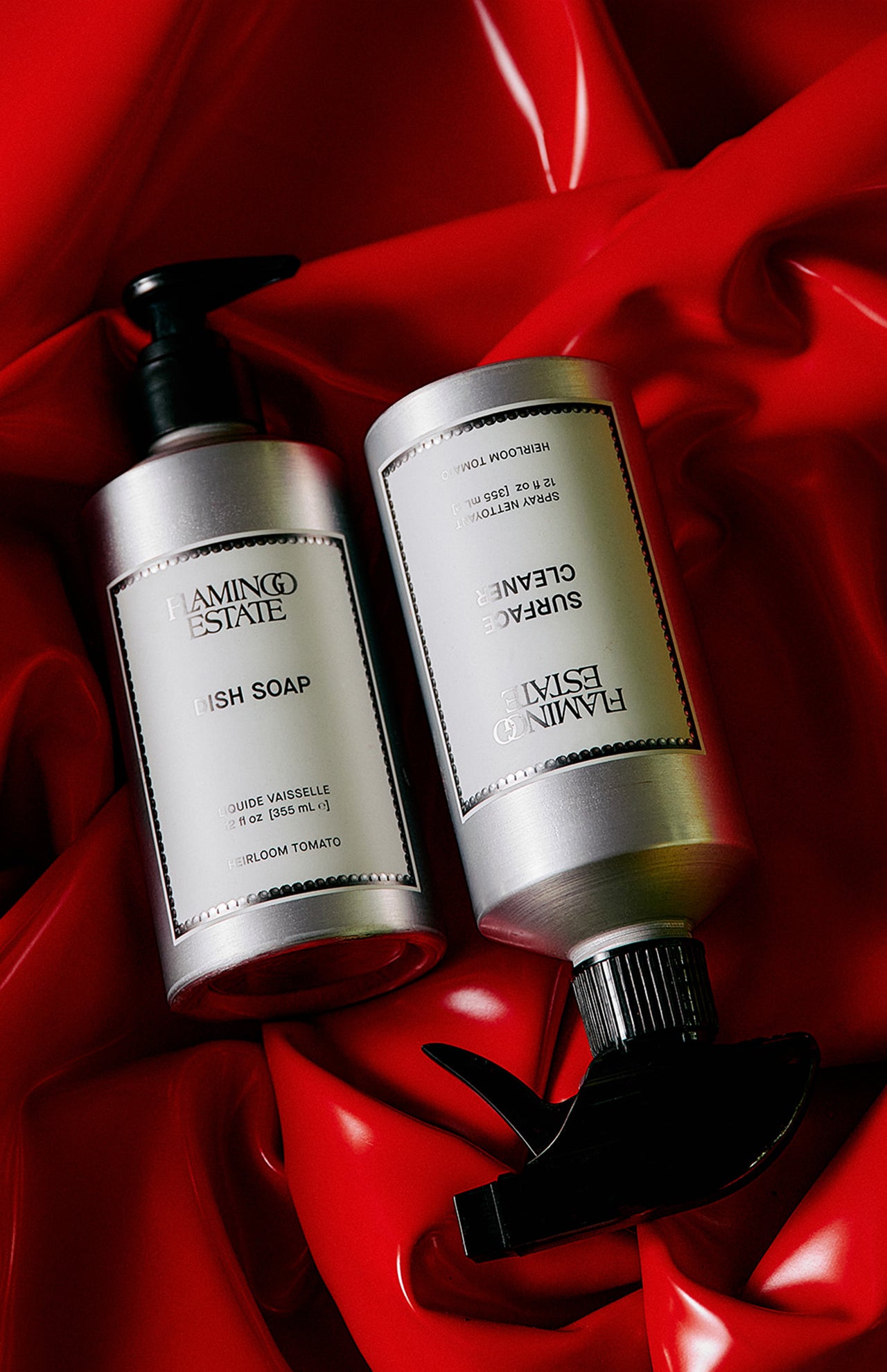 Two silver pump bottles labeled FLAMINGO ESTATE Heirloom Tomato Dish Soap rest on glossy red, crumpled fabric. Minimal labels and black pumps suggest the biodegradable cleanser’s signature tomato leaf scent.