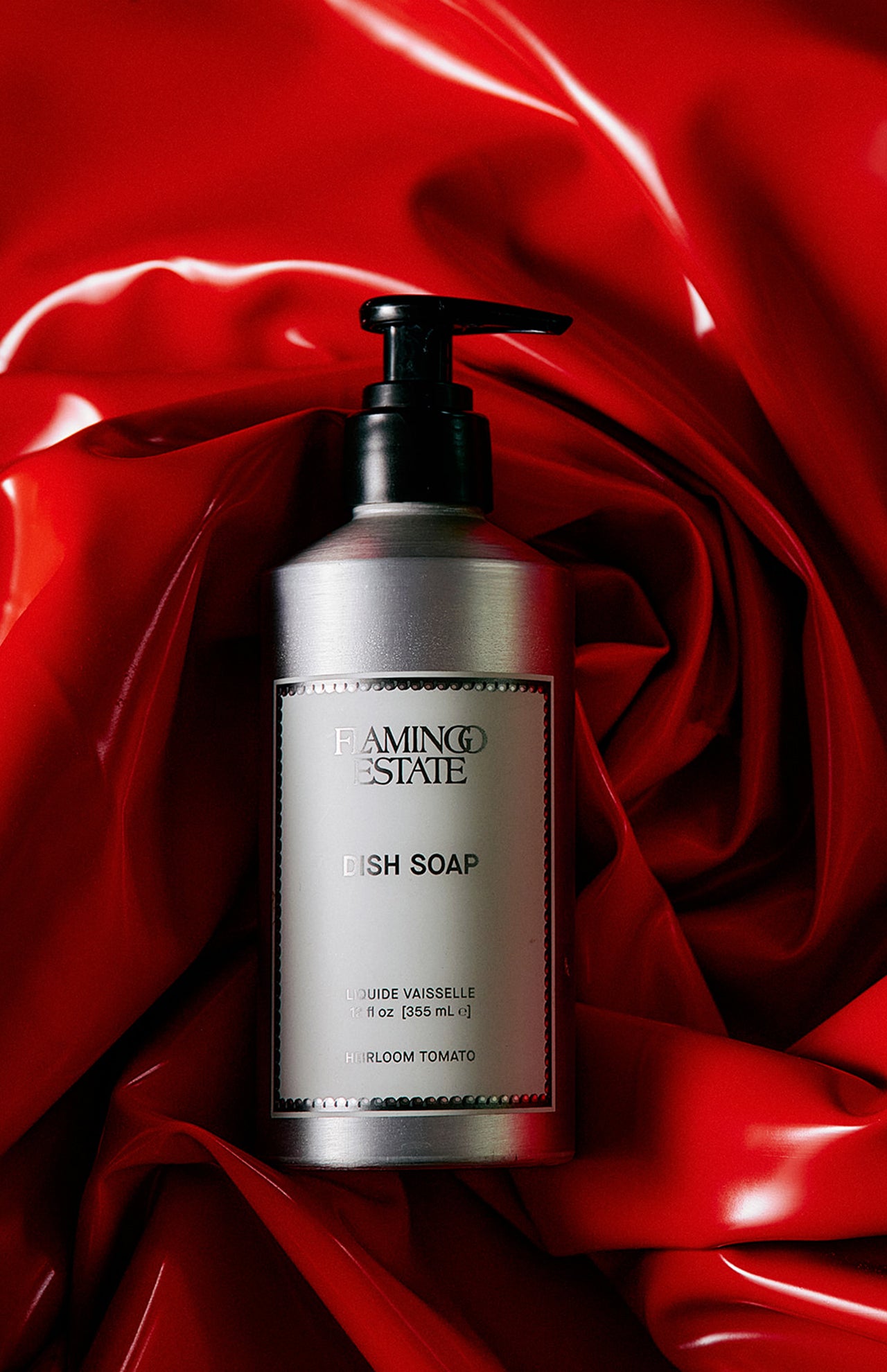 A silver pump bottle of FLAMINGO ESTATE Heirloom Tomato Dish Soap rests on shiny, crumpled red fabric, suggesting a tomato leaf scent and made with biodegradable cleansers for an eco-friendly clean.