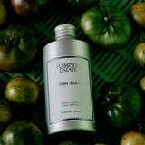 A silver bottle of FLAMINGO ESTATE Heirloom Tomato Dish Soap sits on a green surface among heirloom tomatoes, evoking a fresh tomato leaf aroma.