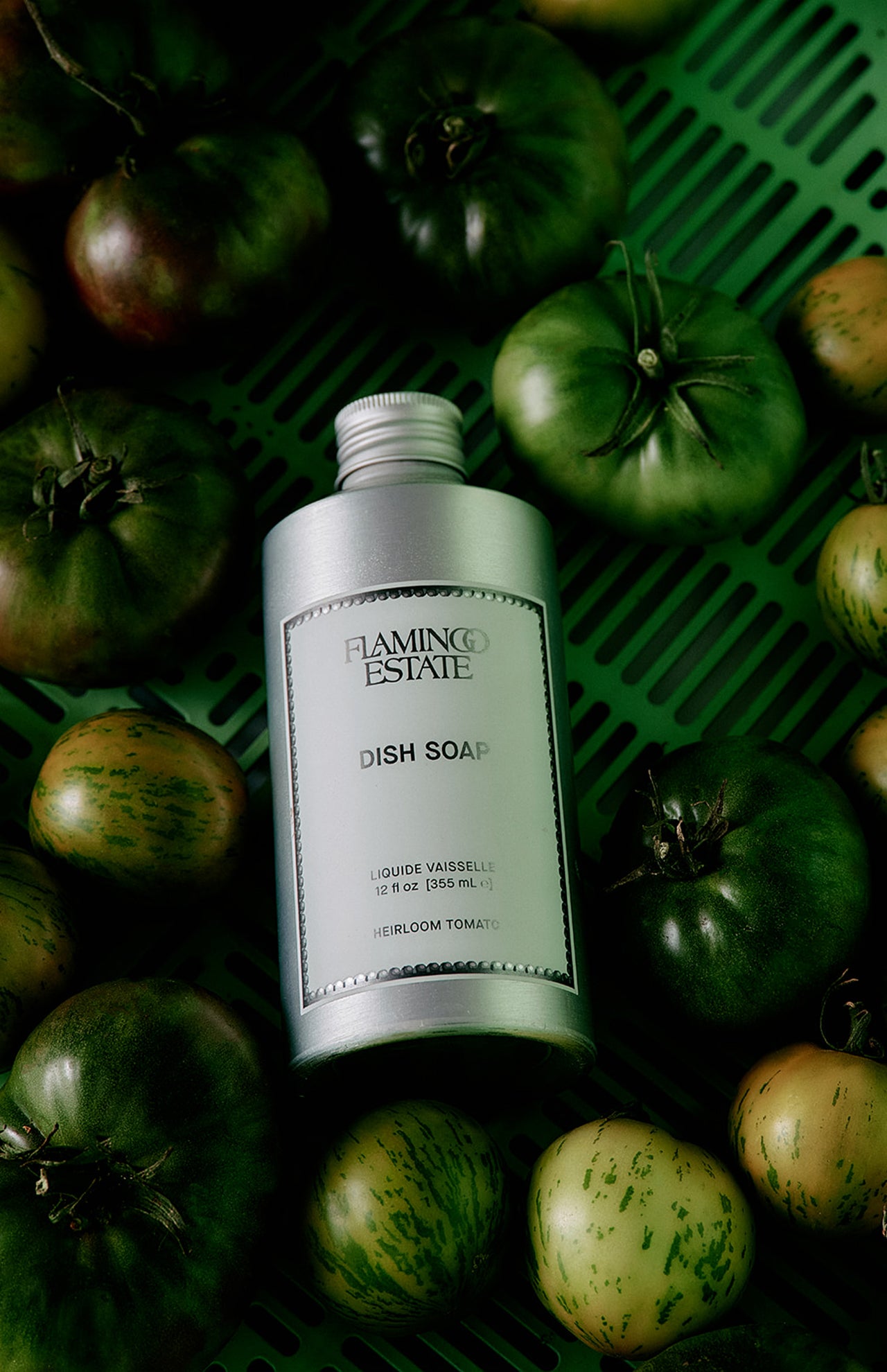 A silver bottle of FLAMINGO ESTATE Heirloom Tomato Dish Soap sits on a green surface among heirloom tomatoes, evoking a fresh tomato leaf aroma.