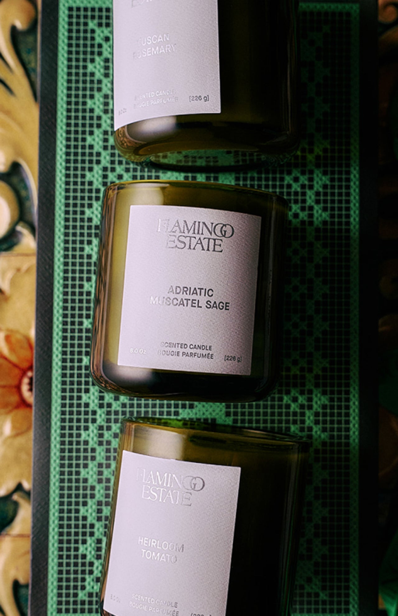 The FLAMINGO ESTATE Three Sisters Candle Set, featuring Tuscan Rosemary, Adriatic Muscatel Sage, and Heirloom Tomato candles, sits on a green grid-patterned tray—an elegant housewarming gift for any home.