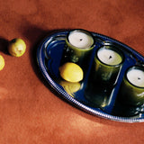 A silver tray holds FLAMINGO ESTATE’s Three Sisters Candle Set—three green candles and a lemon—on a textured brown surface, with two small lemons nearby, making this set an ideal housewarming gift or centerpiece.