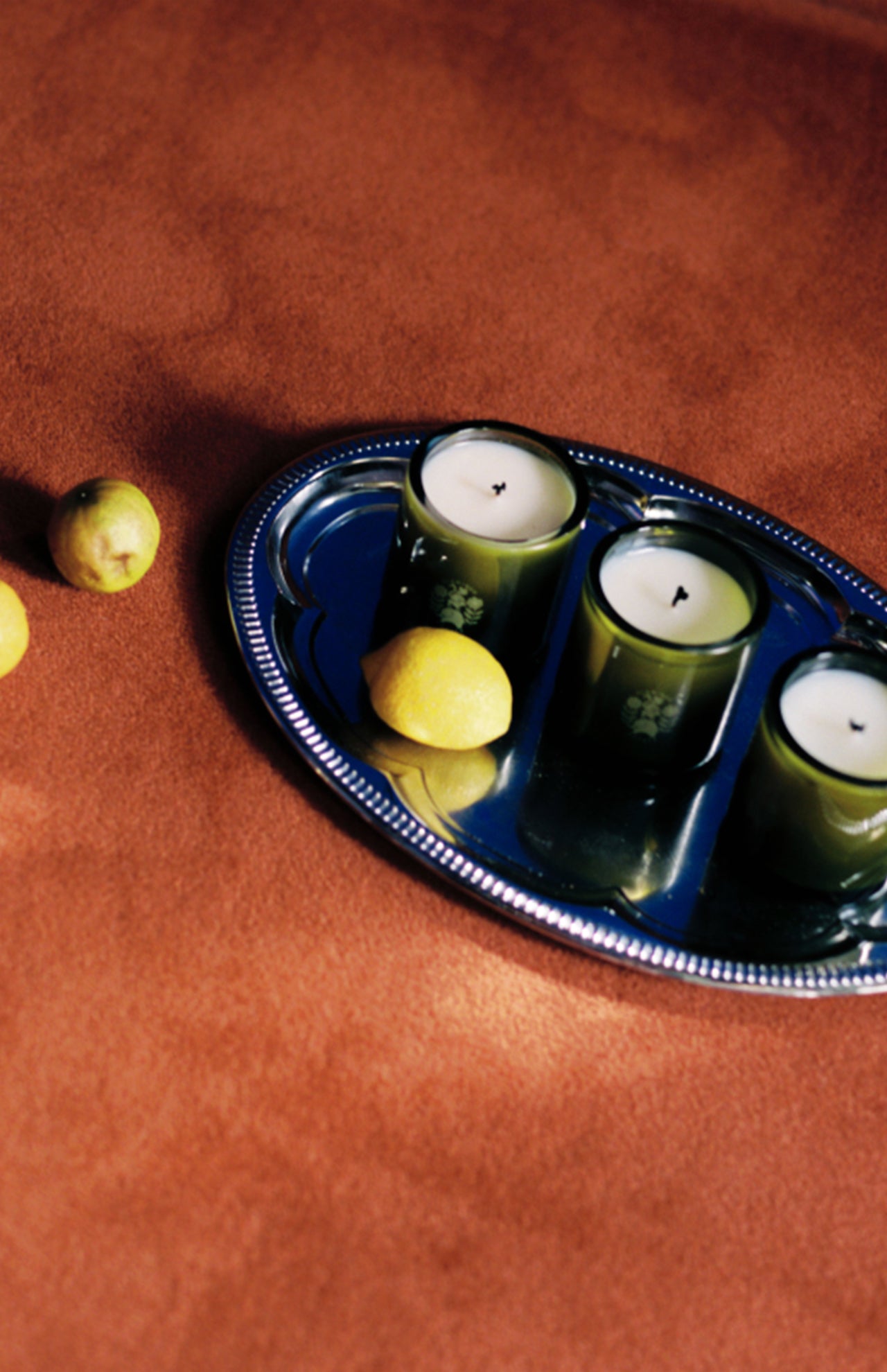 A silver tray holds FLAMINGO ESTATE’s Three Sisters Candle Set—three green candles and a lemon—on a textured brown surface, with two small lemons nearby, making this set an ideal housewarming gift or centerpiece.