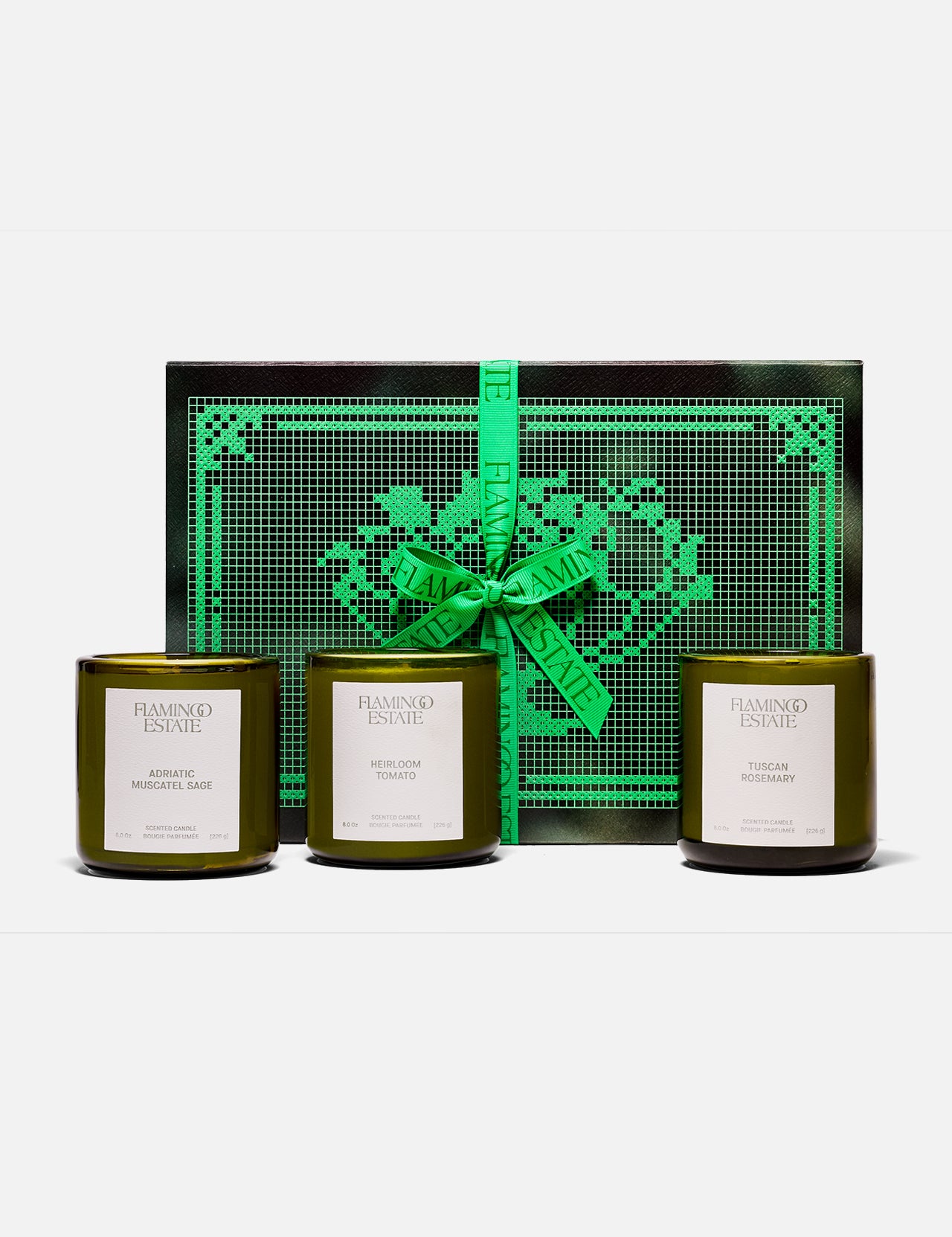 Three green glass FLAMINGO ESTATE Three Sisters candles with white labels sit in front of a black and green heart-patterned box tied with a bright green ribbon—the perfect gift set for a housewarming.