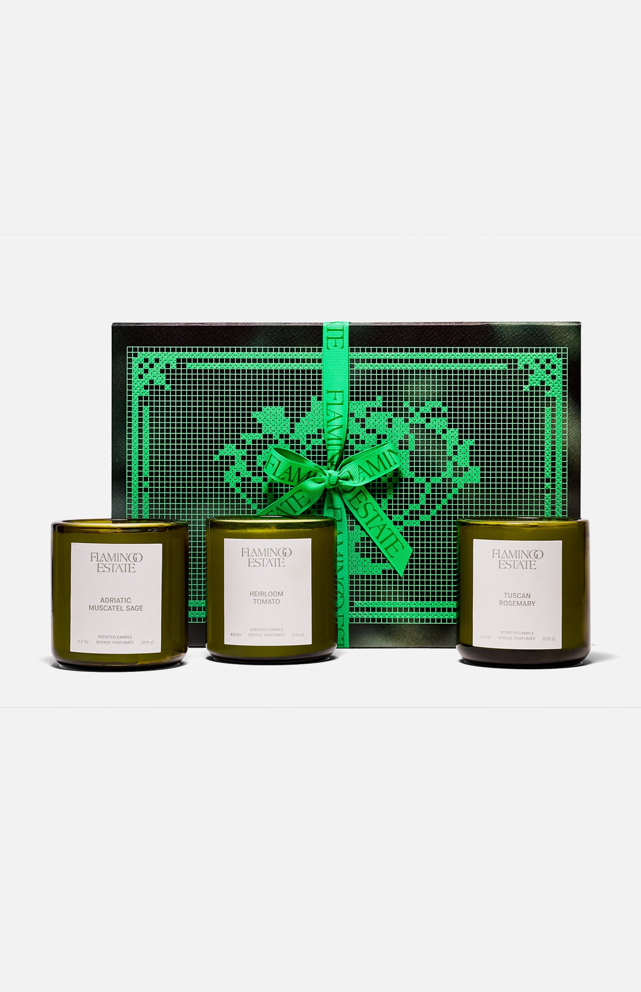 Three green glass FLAMINGO ESTATE Three Sisters candles with white labels sit in front of a black and green heart-patterned box tied with a bright green ribbon—the perfect gift set for a housewarming.