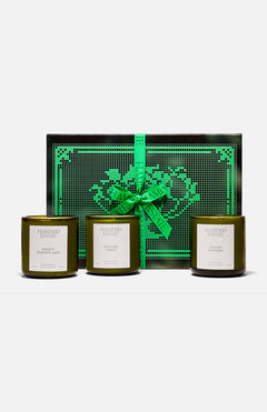 Three green glass FLAMINGO ESTATE Three Sisters candles with white labels sit in front of a black and green heart-patterned box tied with a bright green ribbon—the perfect gift set for a housewarming.