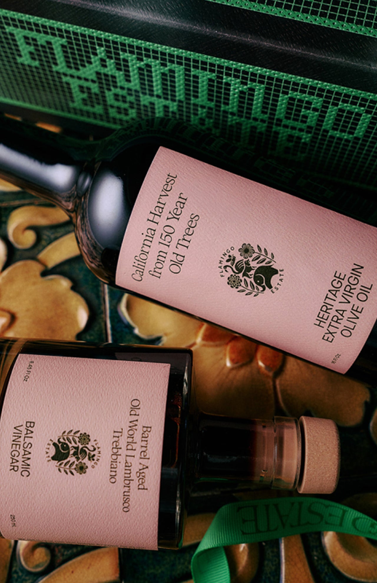 The Tablemates Boxed Oil & Vinegar Gift Set by FLAMINGO ESTATE features two bottles with light pink labels and black text on a decorative surface. Finished with a green ribbon, this elegant set includes Heritage Olive Oil and Balsamic Vinegar.