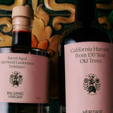 The Tablemates Boxed Oil & Vinegar Gift Set by FLAMINGO ESTATE features two glass bottles with pink labels, elegant fonts, and botanical illustrations, beautifully displayed against a decorative tile backdrop—an ideal charming gift.