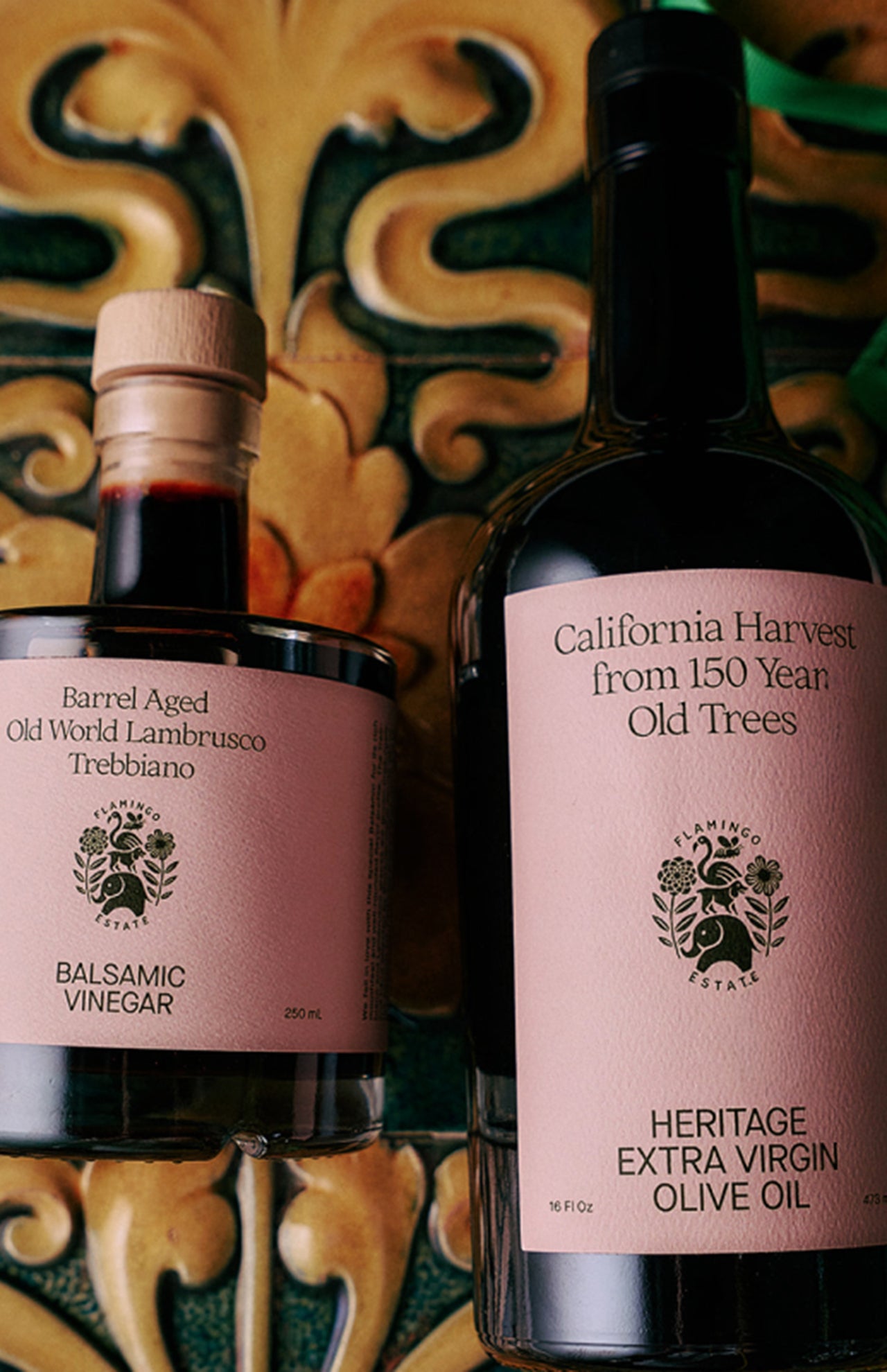 The Tablemates Boxed Oil & Vinegar Gift Set by FLAMINGO ESTATE features two glass bottles with pink labels, elegant fonts, and botanical illustrations, beautifully displayed against a decorative tile backdrop—an ideal charming gift.