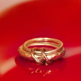 Two yellow gold Agon Vulcan Rings by SPINELLI KILCOLLIN are intertwined on a glossy red surface, casting a striking reflection. The softly blurred background accentuates the brand's elegant modular jewelry design.