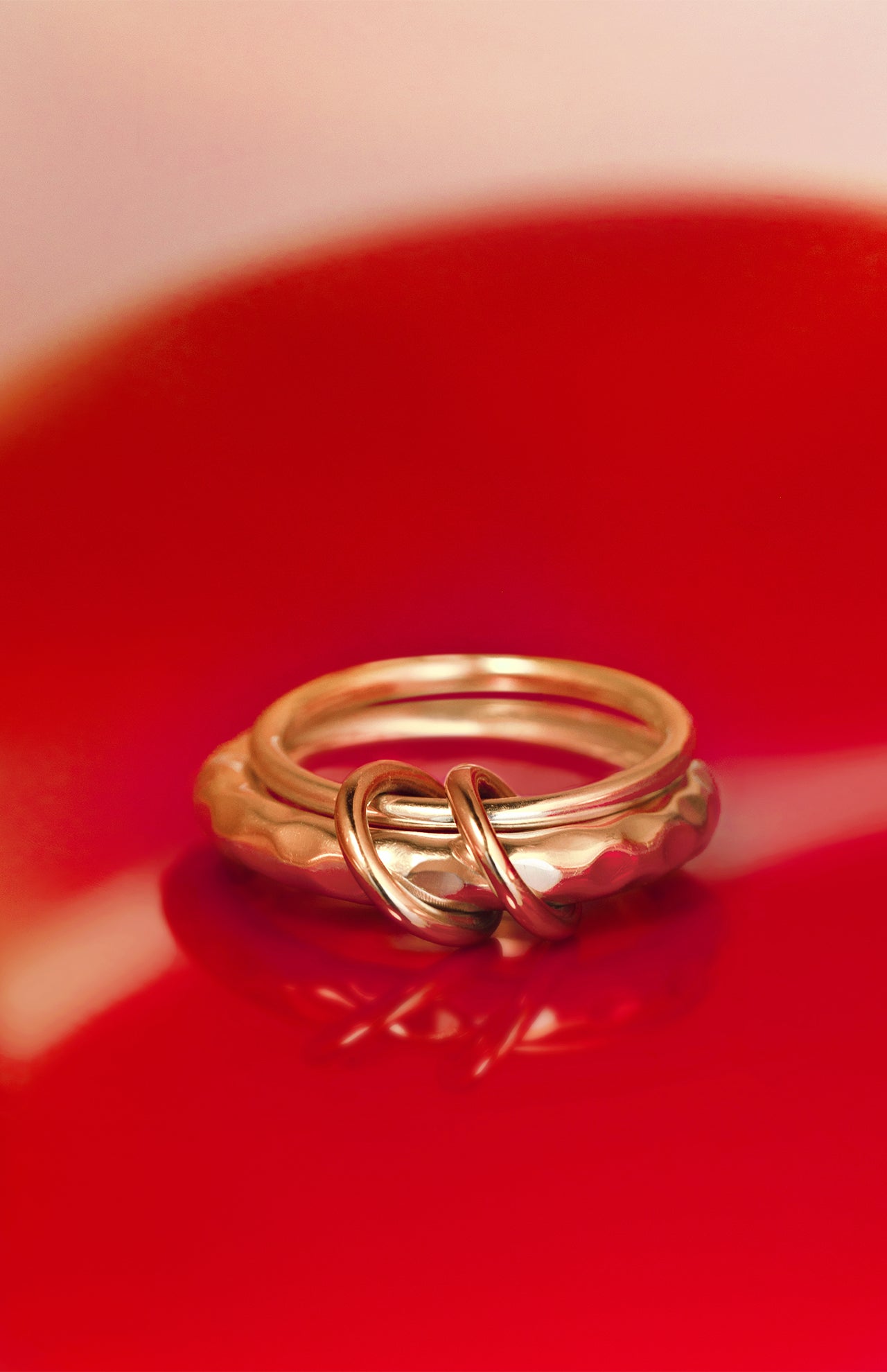 Two yellow gold Agon Vulcan Rings by SPINELLI KILCOLLIN are intertwined on a glossy red surface, casting a striking reflection. The softly blurred background accentuates the brand's elegant modular jewelry design.