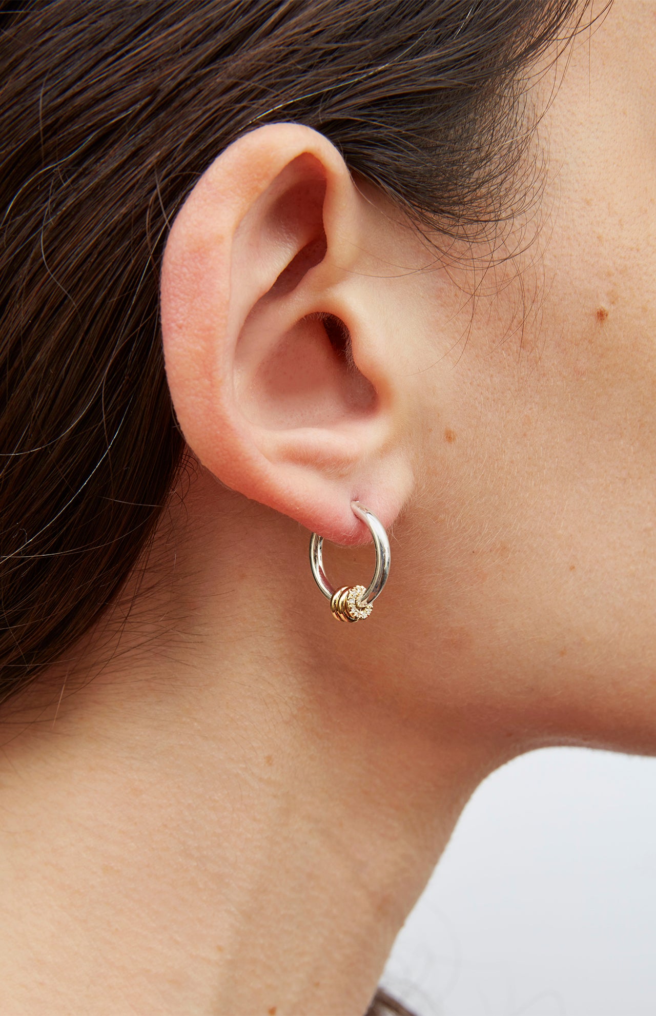 Close-up of a person's ear wearing SPINELLI KILCOLLIN's Ara Deux Diamond Detail Earrings. The sterling silver hoops with gold accents stand out against their straight brown hair and light skin, highlighting the elegant design.