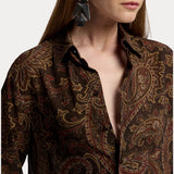 A woman with long hair wears the RALPH LAUREN COLLECTION Paisley Collared Long Sleeve Blouse in a dark brown paisley print, left partly unbuttoned, paired with large geometric black earrings. Shown in partial profile against a white background, this look is an effortlessly stylish choice for any fall wardrobe.