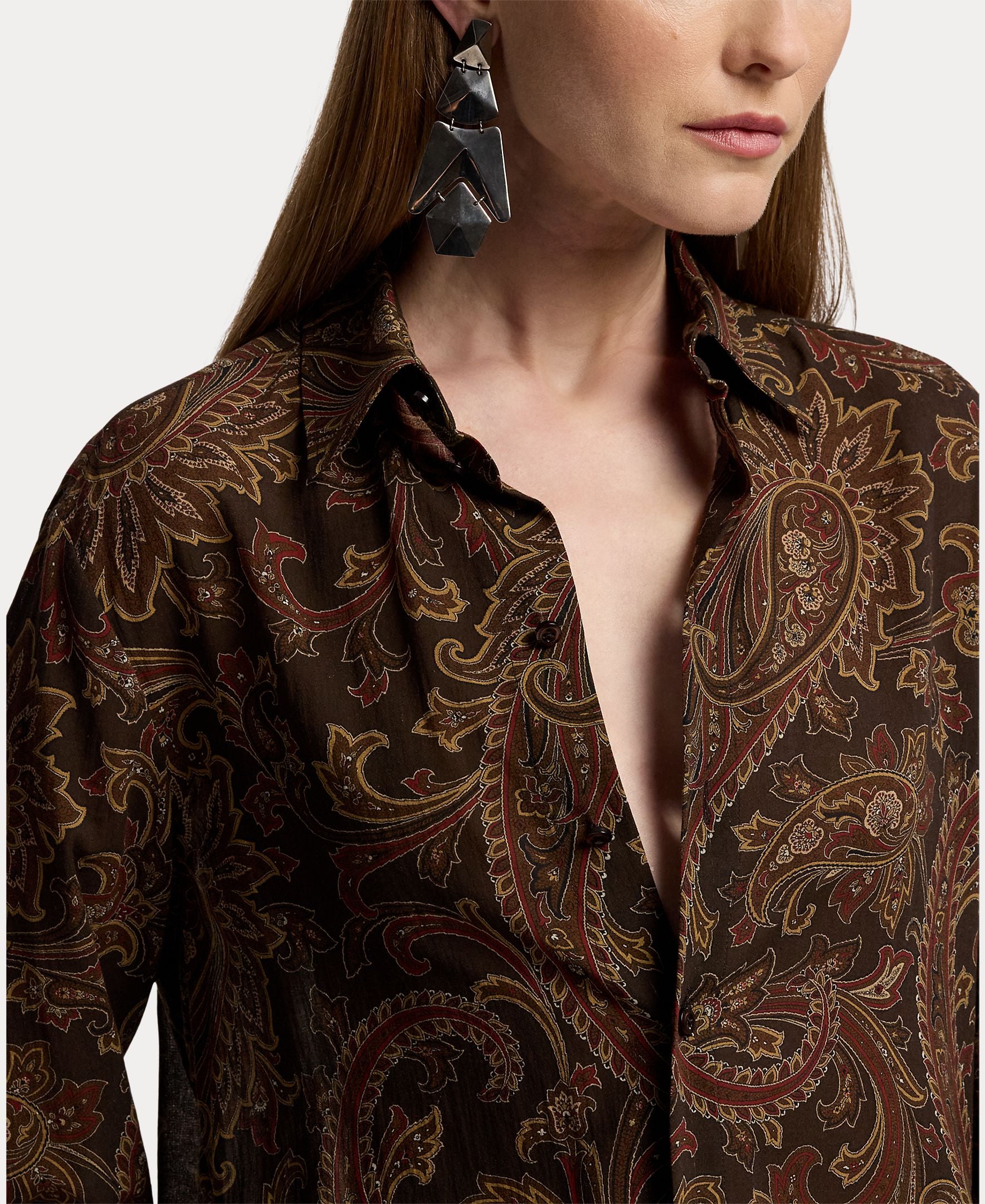 A woman with long hair wears the RALPH LAUREN COLLECTION Paisley Collared Long Sleeve Blouse in a dark brown paisley print, left partly unbuttoned, paired with large geometric black earrings. Shown in partial profile against a white background, this look is an effortlessly stylish choice for any fall wardrobe.