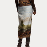 A person is shown from the back wearing the RALPH LAUREN COLLECTION Scenic Silk Twill Pencil Skirt, featuring a striking landscape print with leopard-print borders, made in Italy. The look is paired with a brown long-sleeve top and dark high-heeled boots.