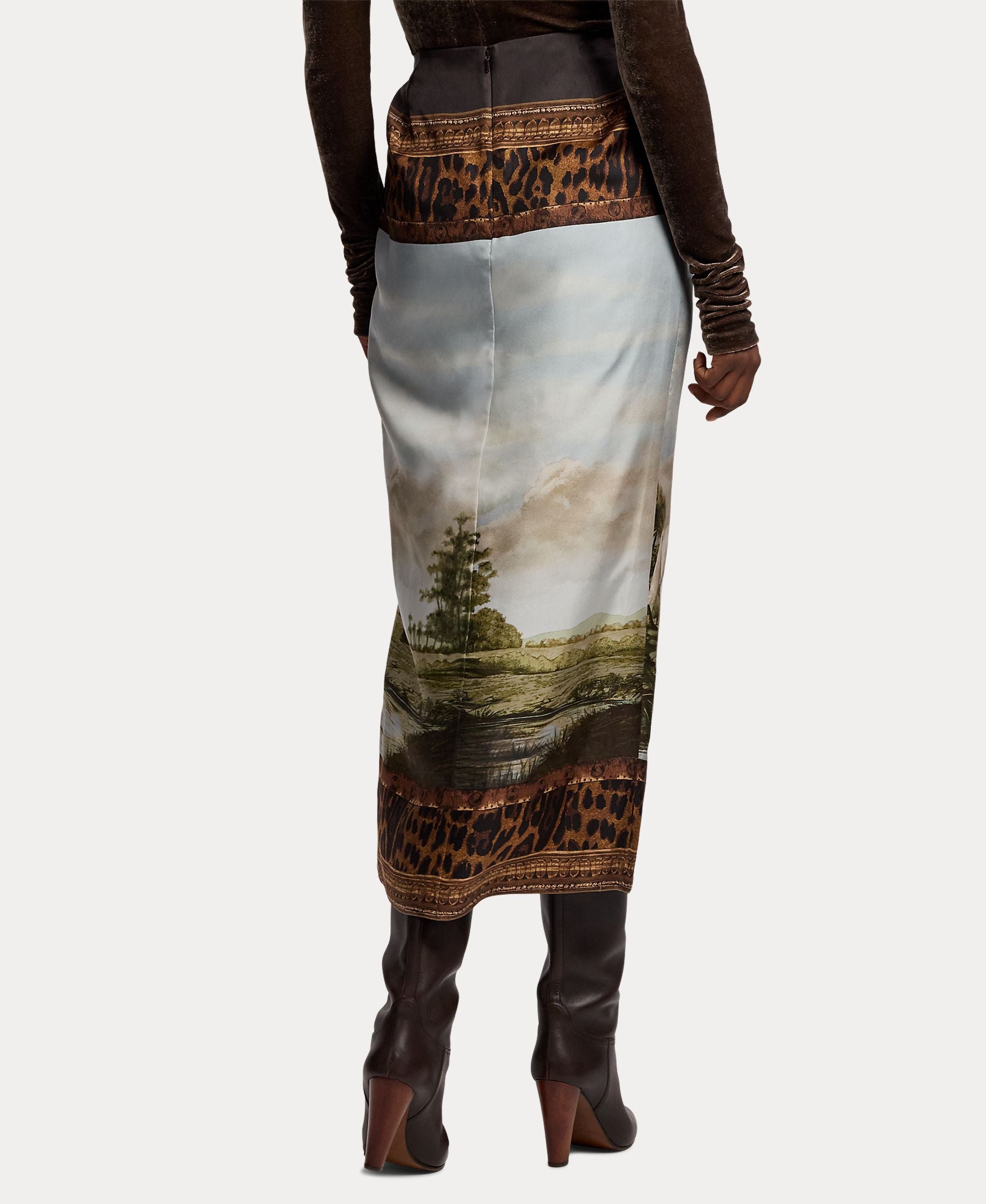 A person is shown from the back wearing the RALPH LAUREN COLLECTION Scenic Silk Twill Pencil Skirt, featuring a striking landscape print with leopard-print borders, made in Italy. The look is paired with a brown long-sleeve top and dark high-heeled boots.