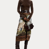 A person wearing a brown velvet turtleneck, the RALPH LAUREN COLLECTION Scenic Silk Twill Pencil Skirt with artistic animal and landscape patterns made in Italy, a wide belt, dark knee-high boots, and holding a small dark handbag.