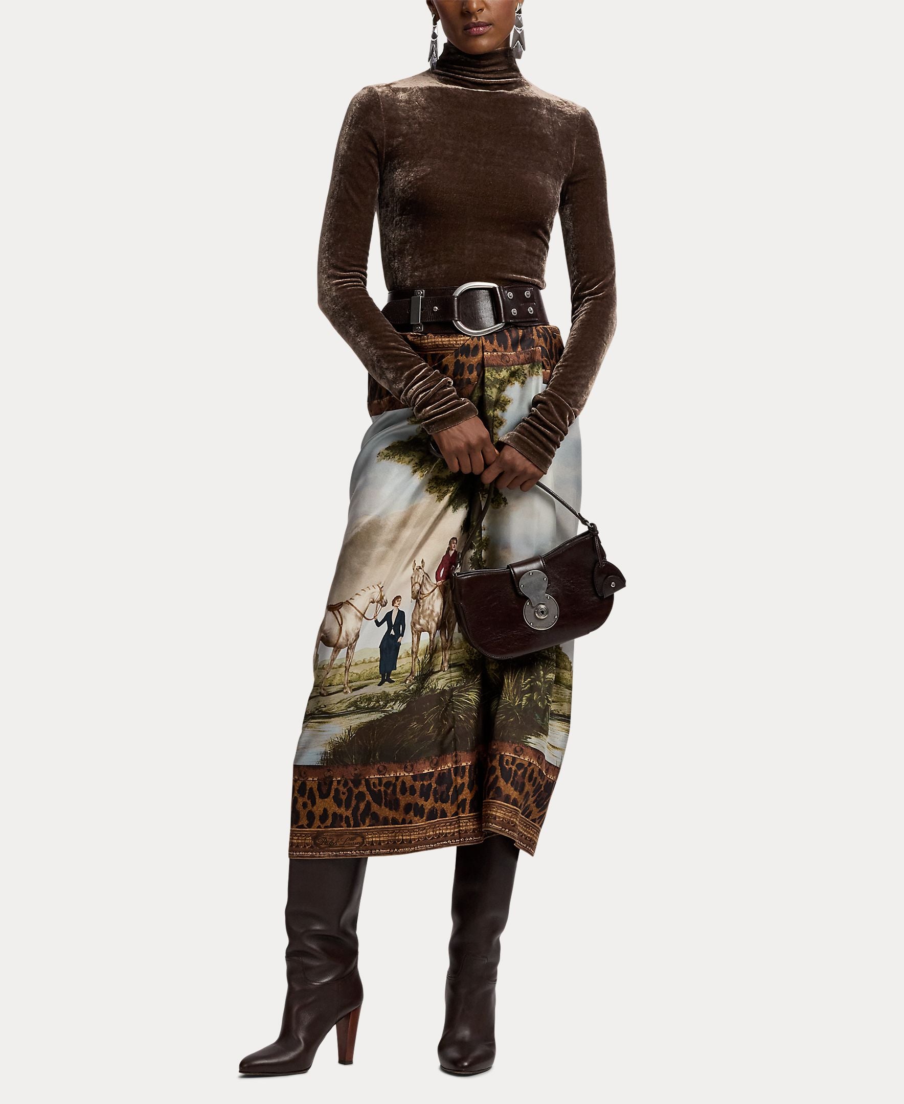 A person wearing a brown velvet turtleneck, the RALPH LAUREN COLLECTION Scenic Silk Twill Pencil Skirt with artistic animal and landscape patterns made in Italy, a wide belt, dark knee-high boots, and holding a small dark handbag.