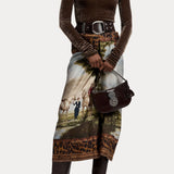 A model wears a brown turtleneck, belt, and Scenic Silk Twill Pencil Skirt featuring a pastoral landscape print with horses and a leopard print border, holding a dark brown handbag.