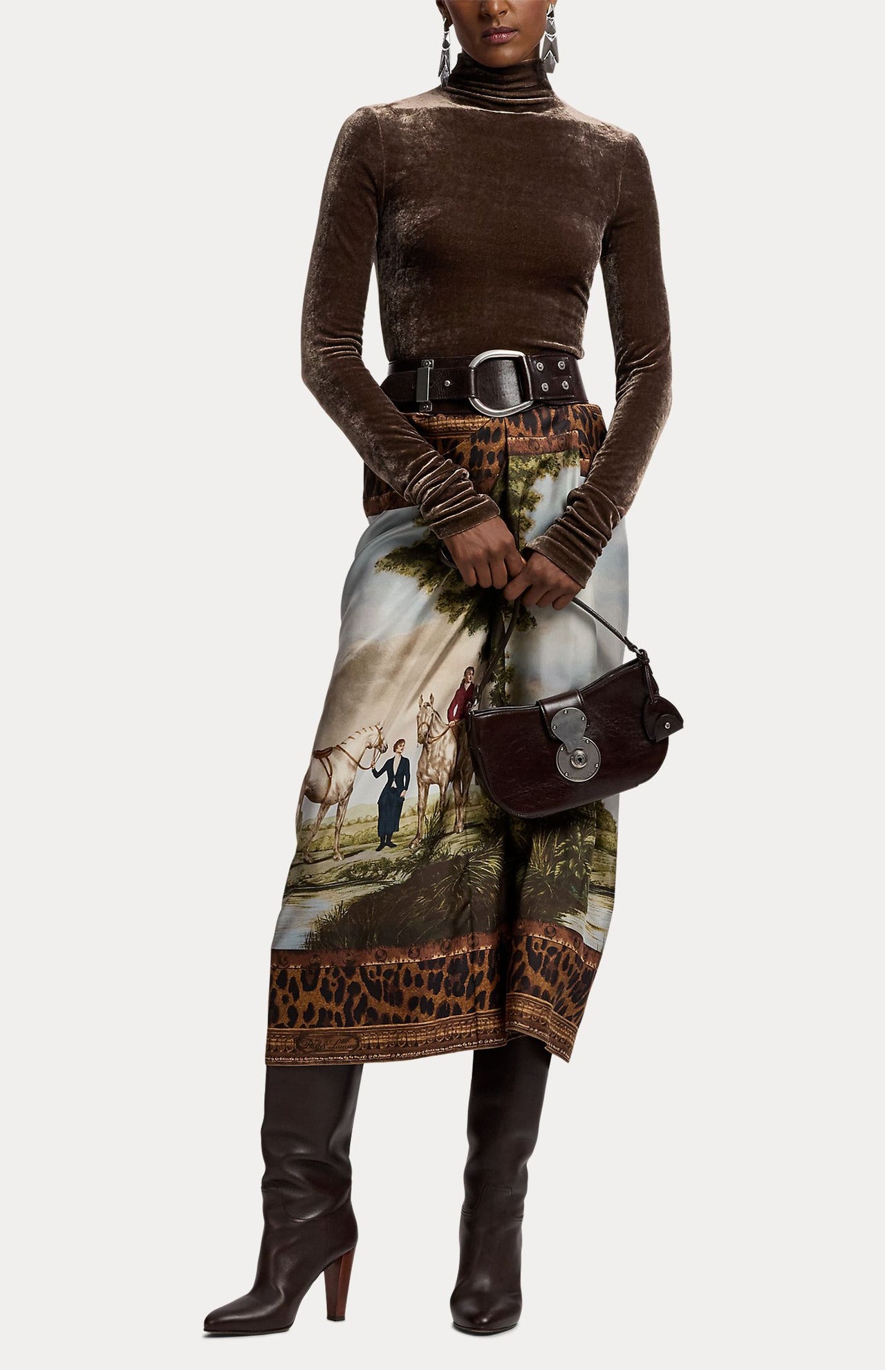 A model wears a brown turtleneck, belt, and Scenic Silk Twill Pencil Skirt featuring a pastoral landscape print with horses and a leopard print border, holding a dark brown handbag.