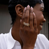 A woman with short hair smiles, holding her manicured hand to her face and showcasing the chunky gold SPINELLI KILCOLLIN Tigris Pavé Band Ring. She wears a white collared shirt and a small gold stud earring.
