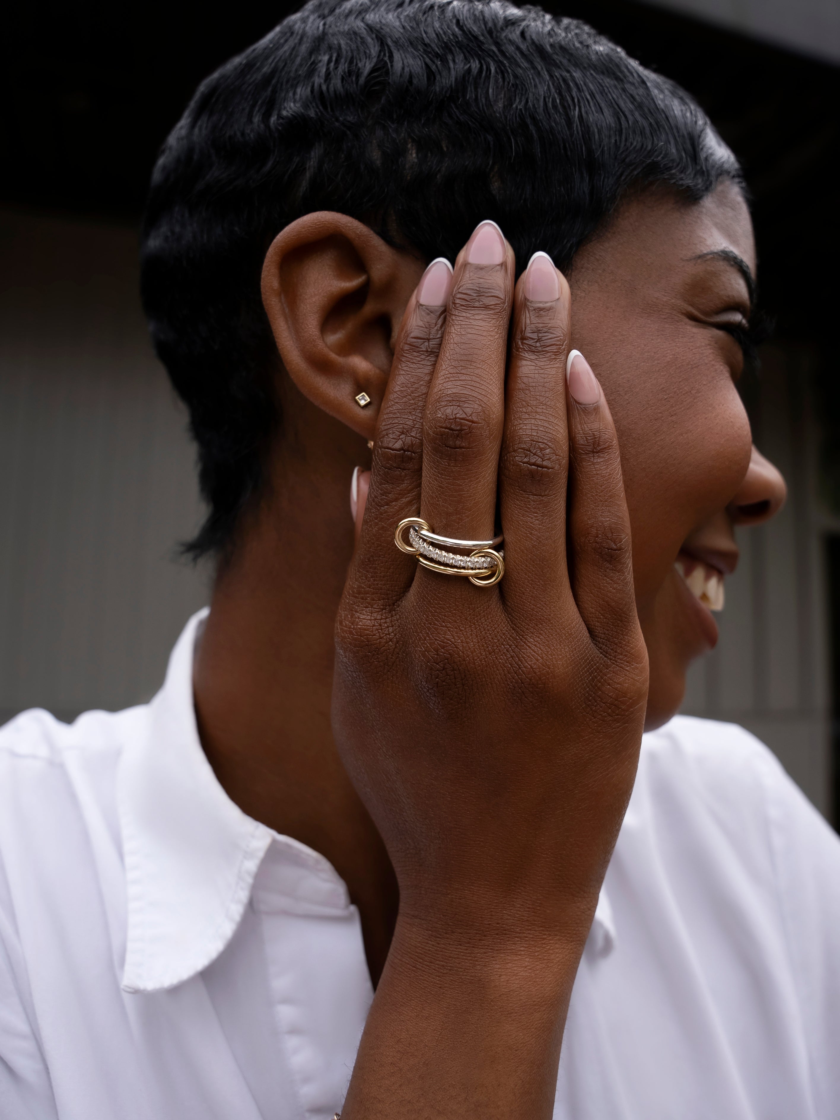 A woman with short hair smiles, holding her manicured hand to her face and showcasing the chunky gold SPINELLI KILCOLLIN Tigris Pavé Band Ring. She wears a white collared shirt and a small gold stud earring.