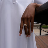 A person wearing a white long-sleeve garment rests their hand on a railing, showcasing the SPINELLI KILCOLLIN Solarium 3 Band Ring with its fluid gold design. Their nails are manicured, and the background features softly blurred outdoor elements.