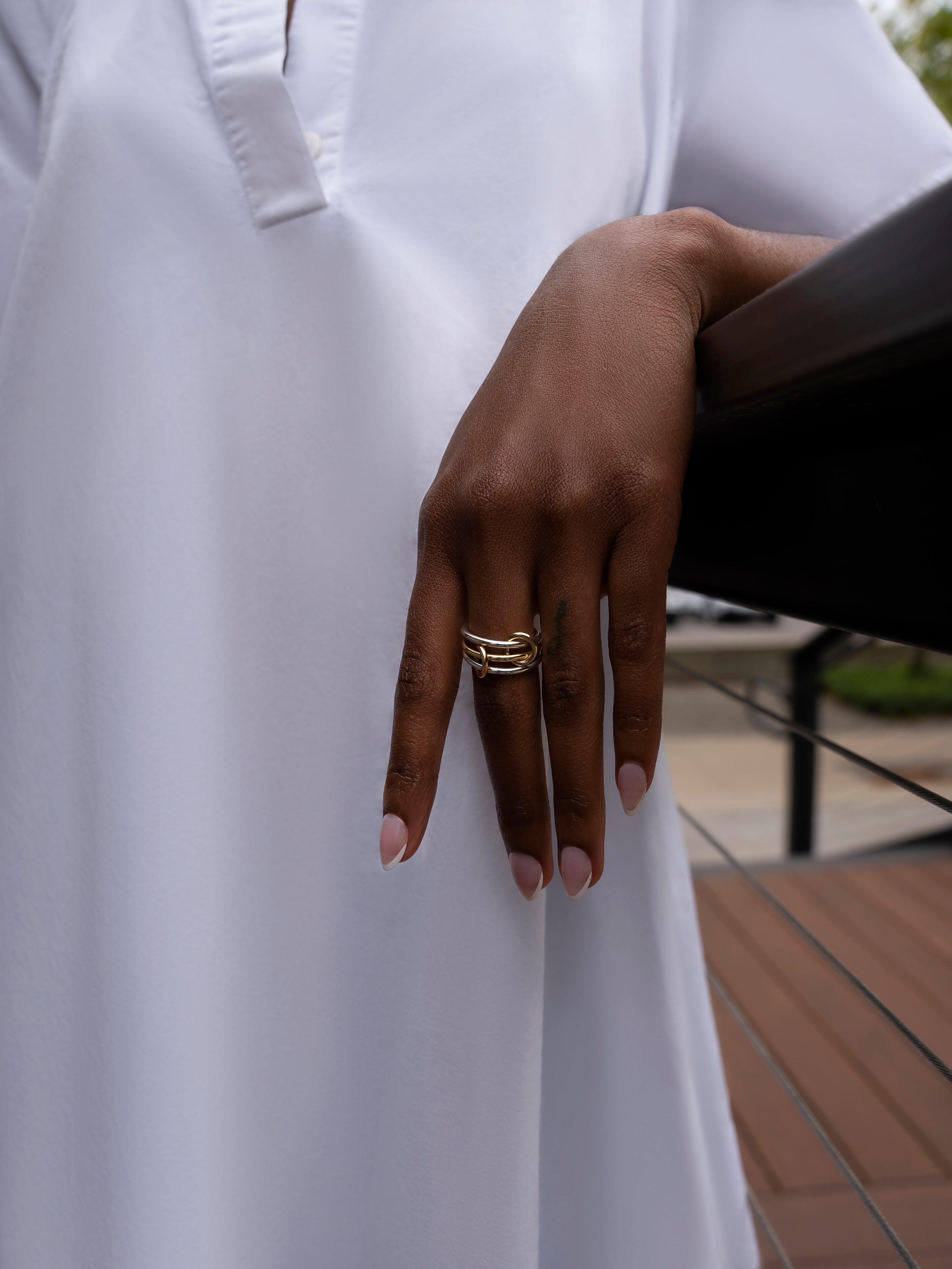 A person wearing a white long-sleeve garment rests their hand on a railing, showcasing the SPINELLI KILCOLLIN Solarium 3 Band Ring with its fluid gold design. Their nails are manicured, and the background features softly blurred outdoor elements.