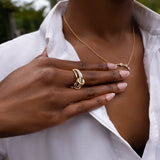A person wearing a white shirt touches their chest, showcasing a gold necklace and the stunning SPINELLI KILCOLLIN Tigris Pavé Band Ring with its signature loop design. Their manicured nails and the softly blurred greenery in the background enhance the elegant allure of this modular jewelry.