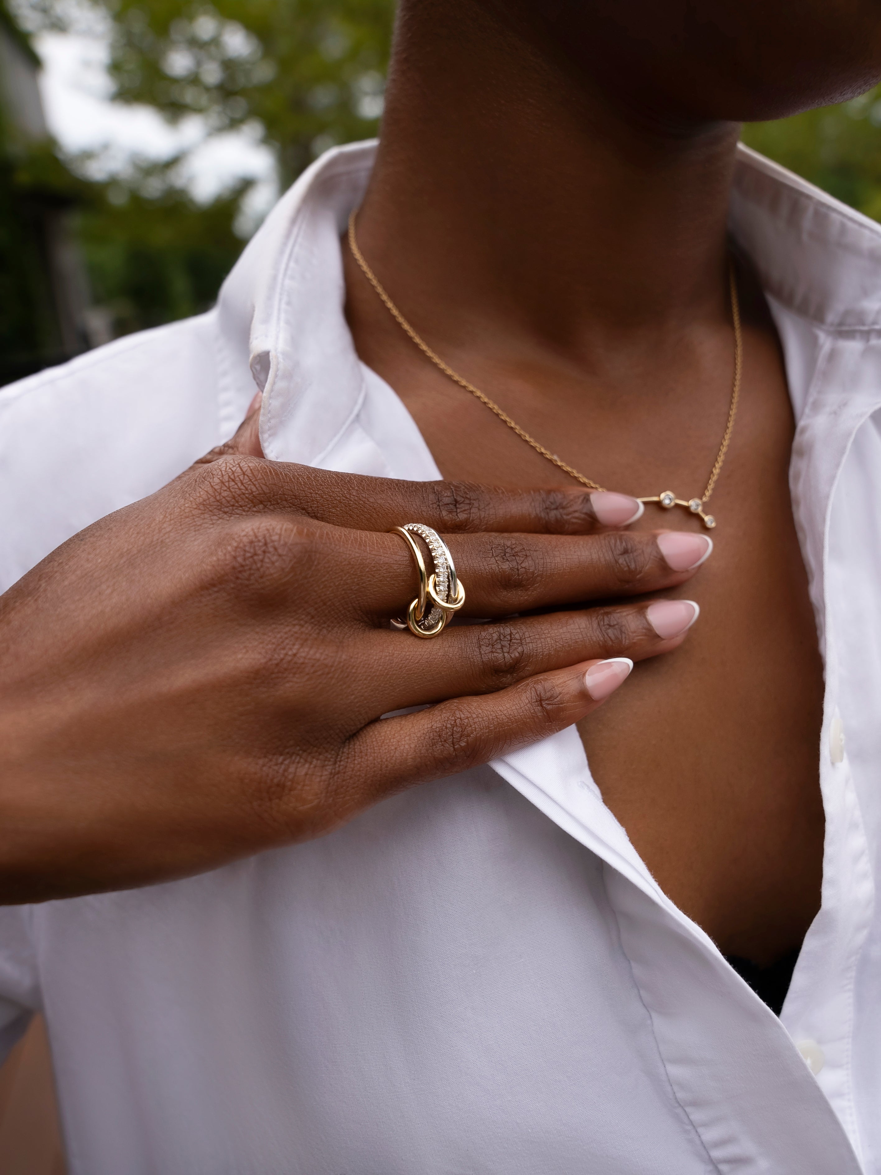 A person wearing a white shirt touches their chest, showcasing a gold necklace and the stunning SPINELLI KILCOLLIN Tigris Pavé Band Ring with its signature loop design. Their manicured nails and the softly blurred greenery in the background enhance the elegant allure of this modular jewelry.