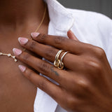A person with manicured nails wears the SPINELLI KILCOLLIN Solarium 3 Band Ring—featuring fluid, interlinked gold bands—on their finger and a delicate gold necklace, dressed in a white button-down shirt.