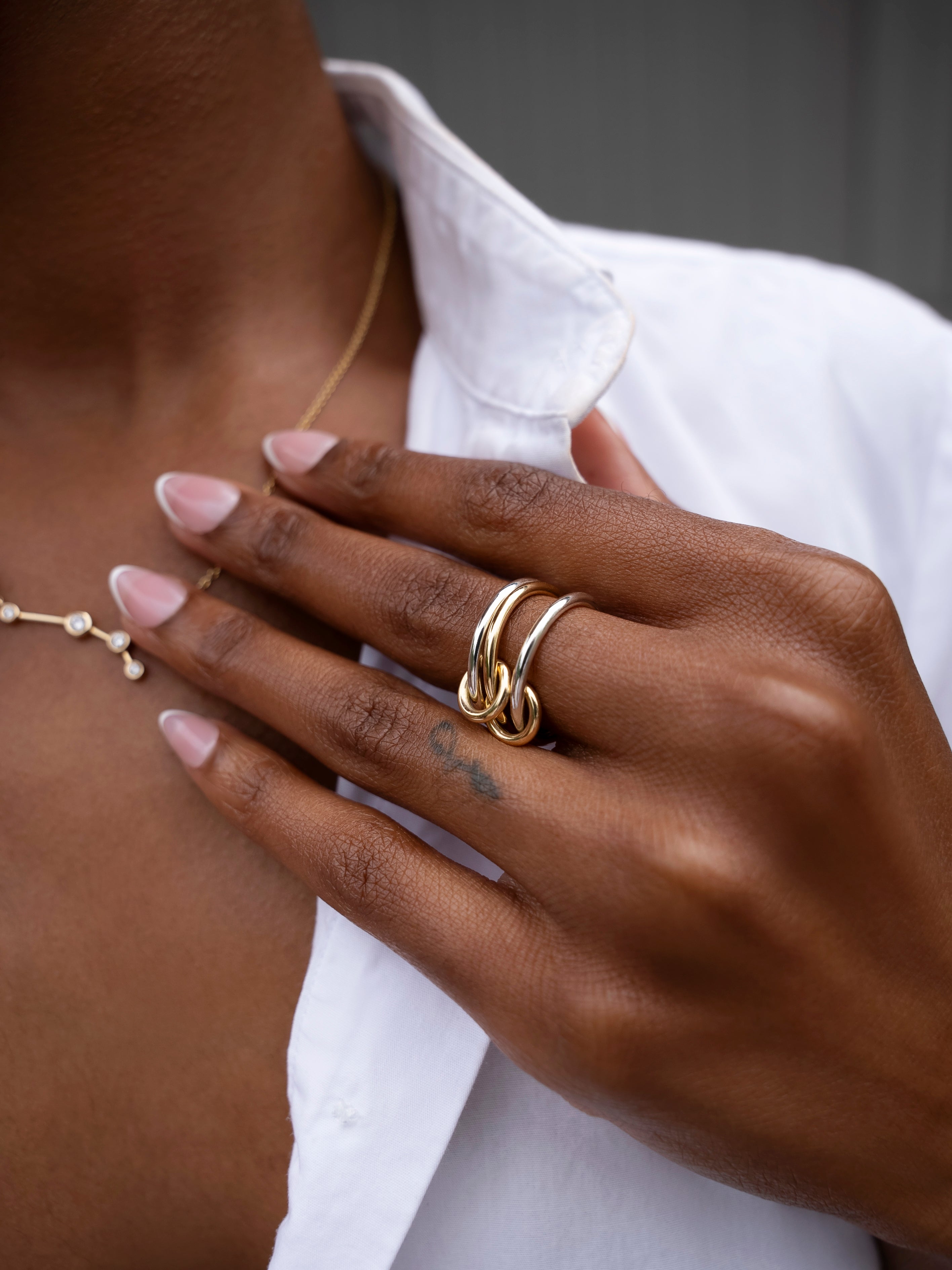 A person with manicured nails wears the SPINELLI KILCOLLIN Solarium 3 Band Ring—featuring fluid, interlinked gold bands—on their finger and a delicate gold necklace, dressed in a white button-down shirt.