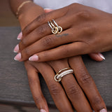 Close-up of hands with French manicured nails, wearing two modular SPINELLI KILCOLLIN Solarium 3 Band Rings—one featuring a yellow gold double loop design and the other accented with a row of small stones—resting on a gray wooden surface.