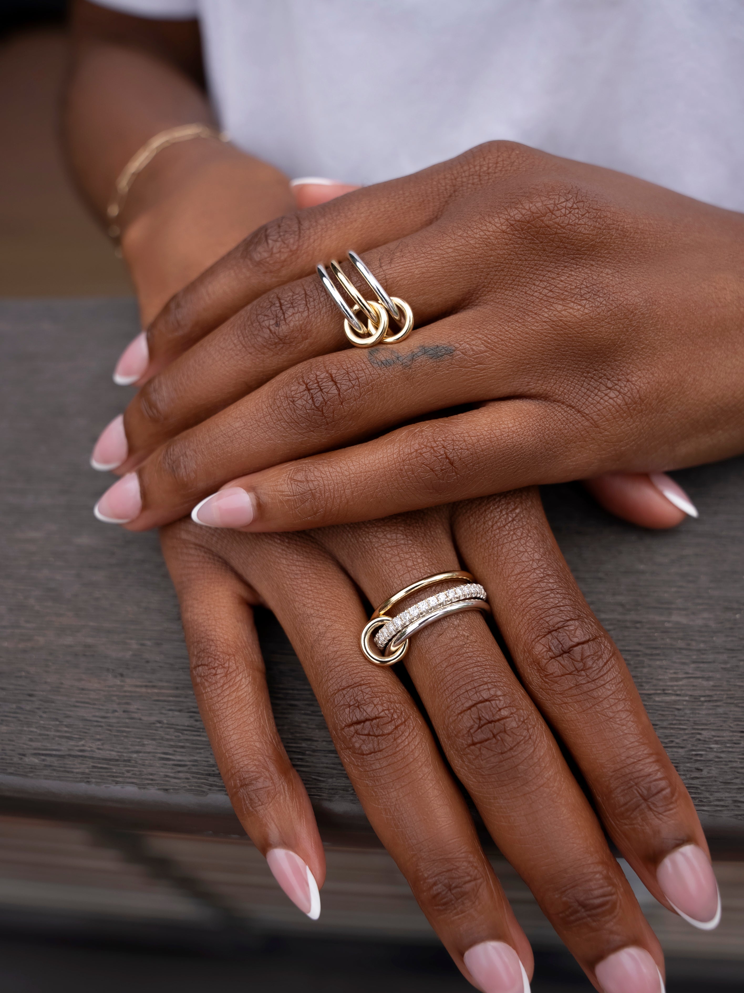 Close-up of hands with French manicured nails, wearing two modular SPINELLI KILCOLLIN Solarium 3 Band Rings—one featuring a yellow gold double loop design and the other accented with a row of small stones—resting on a gray wooden surface.