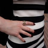 A hand adorned with deep red nails and multiple rings, including the SPINELLI KILCOLLIN Nimbus 4 Band Diamond Ring, rests atop a black and white striped dress against a backdrop of wooden flooring.