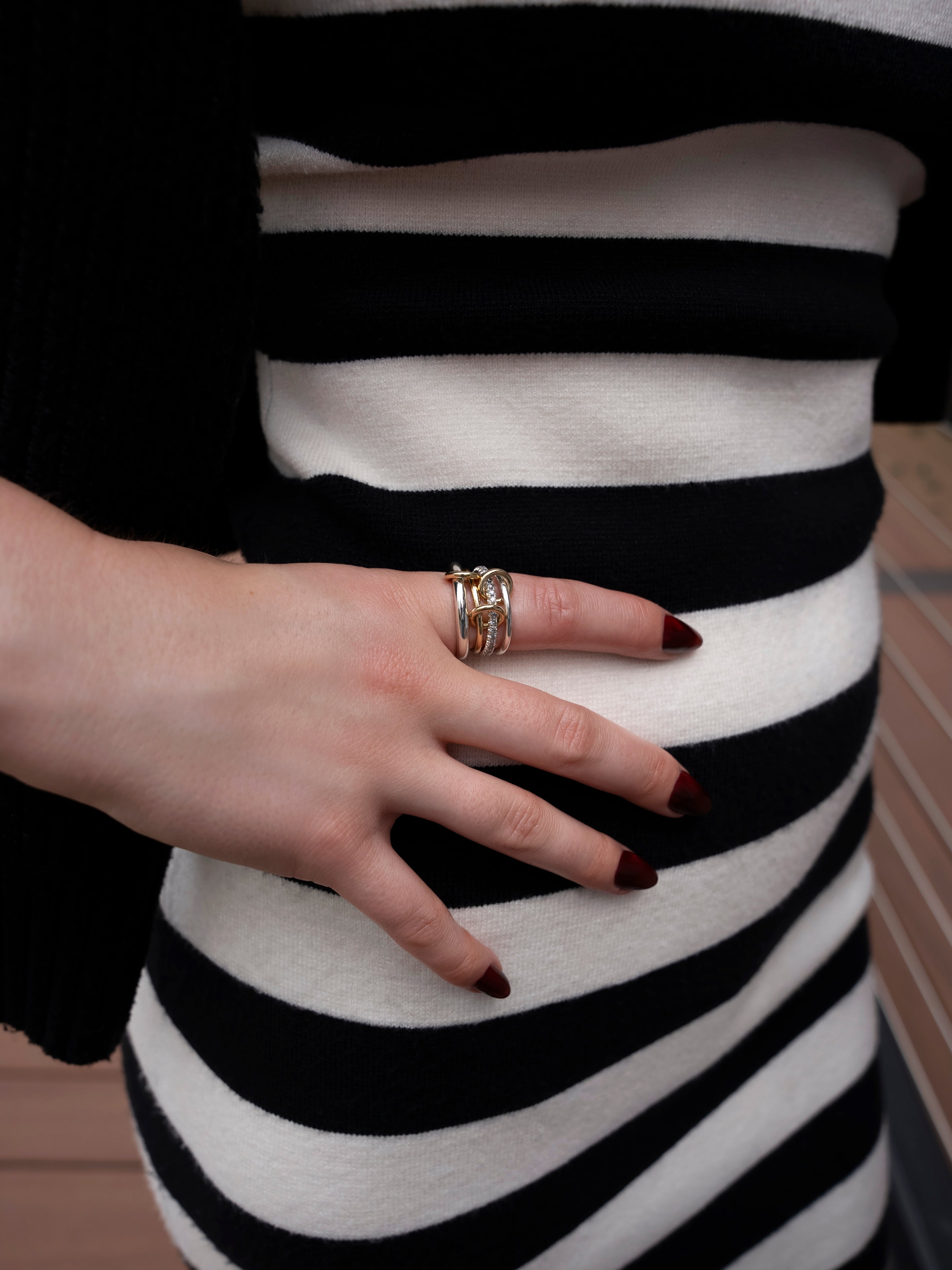 A hand adorned with deep red nails and multiple rings, including the SPINELLI KILCOLLIN Nimbus 4 Band Diamond Ring, rests atop a black and white striped dress against a backdrop of wooden flooring.