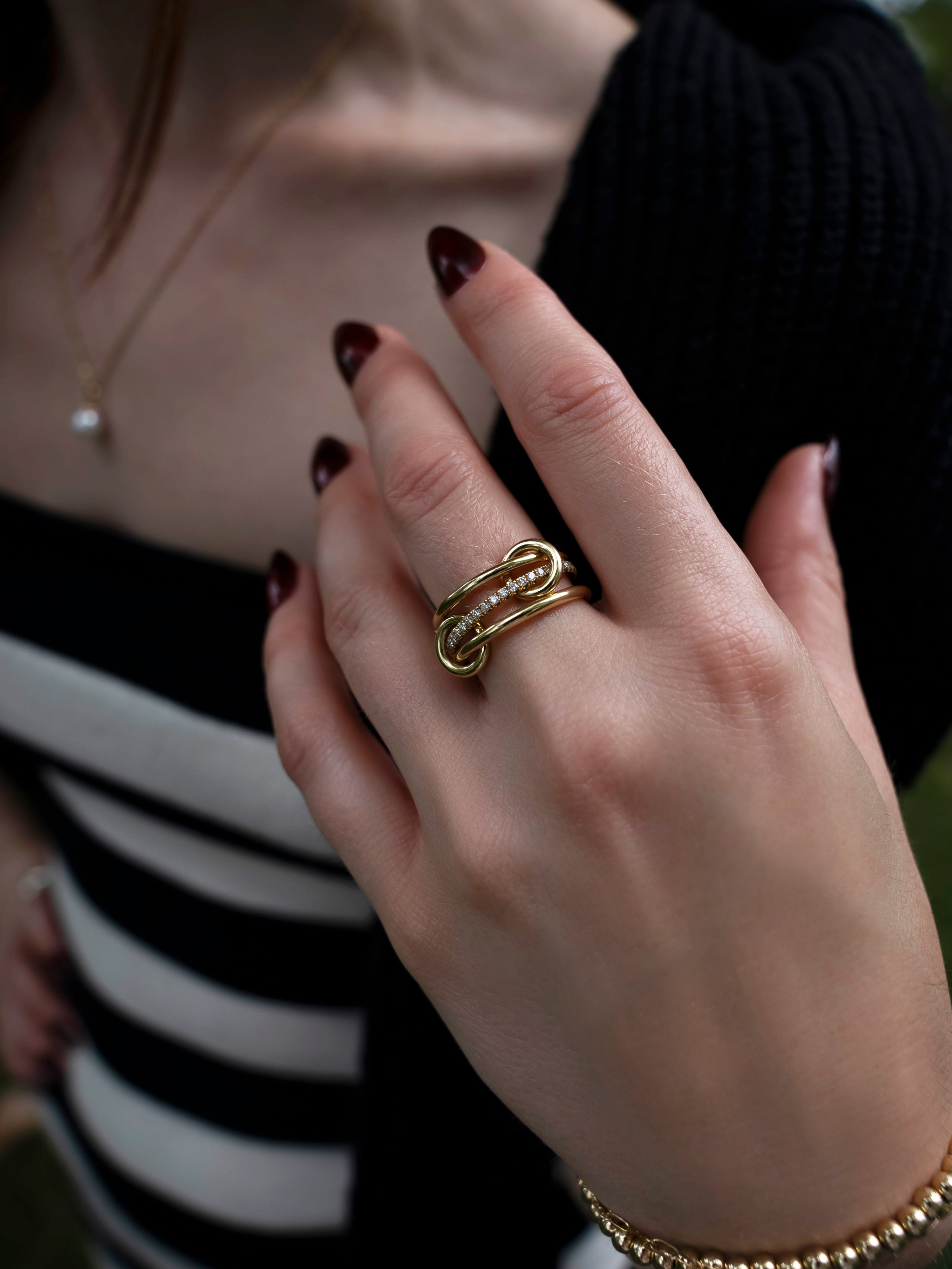 A close-up of a woman’s hand wearing the SPINELLI KILCOLLIN Sonny Pavé Band Ring in yellow gold, featuring a unique modular design, paired with dark red nail polish and a gold bracelet. She is dressed in a black and white striped top and a black sweater.