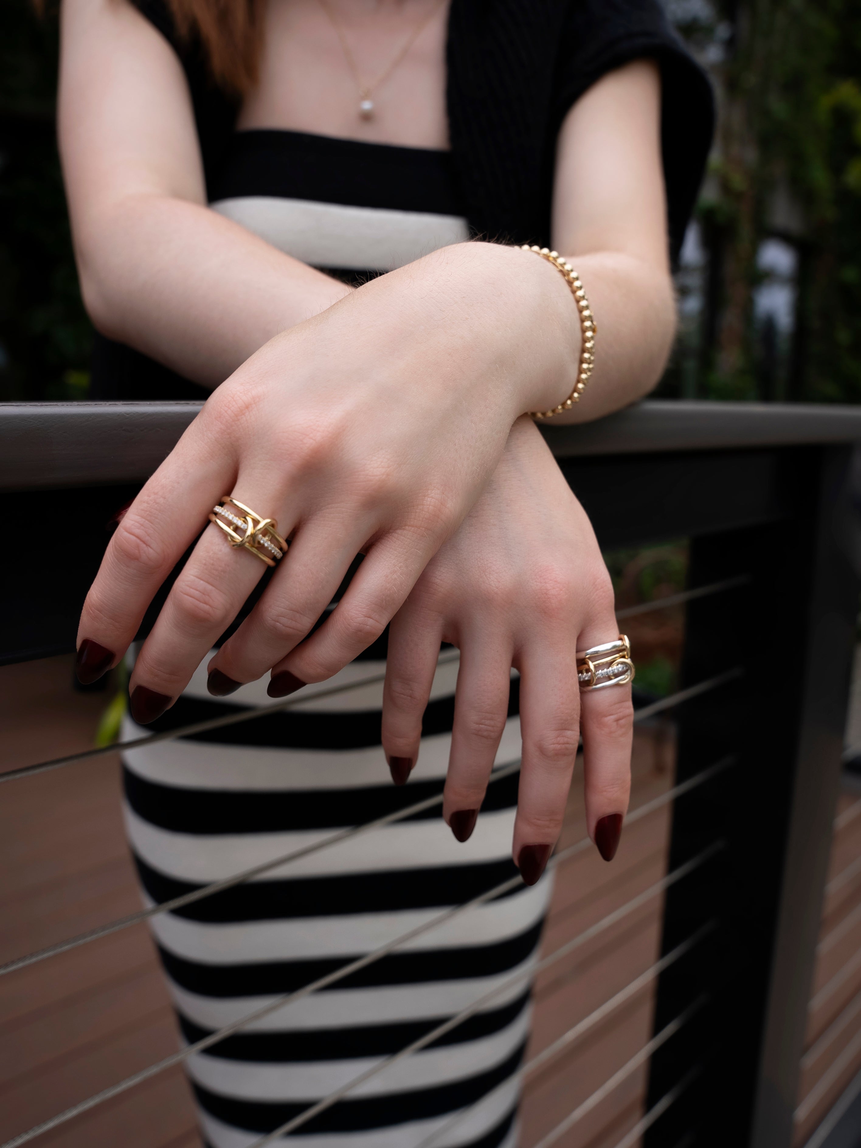 A person with dark nail polish and gold jewelry, including the Nimbus 4 Band Diamond Ring by SPINELLI KILCOLLIN, rests their hands on a railing while wearing a black and white striped dress.