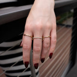 A hand with dark red nails wears the Sonny Pavé Band Ring by SPINELLI KILCOLLIN, along with a gold bracelet and another gold ring set with small pearls, resting on a metal railing. The background is blurred, revealing part of a striped black-and-white outfit.
