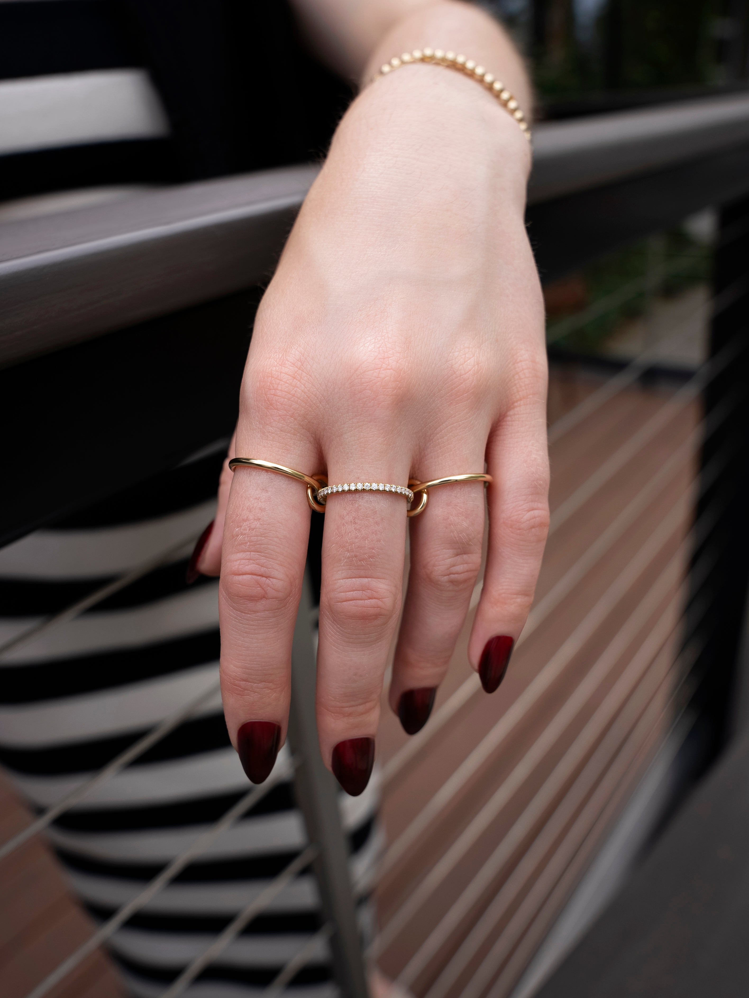 A hand with dark red nails wears the Sonny Pavé Band Ring by SPINELLI KILCOLLIN, along with a gold bracelet and another gold ring set with small pearls, resting on a metal railing. The background is blurred, revealing part of a striped black-and-white outfit.