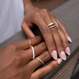 A close-up of hands with a light pink French manicure resting on a railing, adorned with multiple gold and pavé diamond rings, including the stunning Tigris Pavé Band Ring by SPINELLI KILCOLLIN.