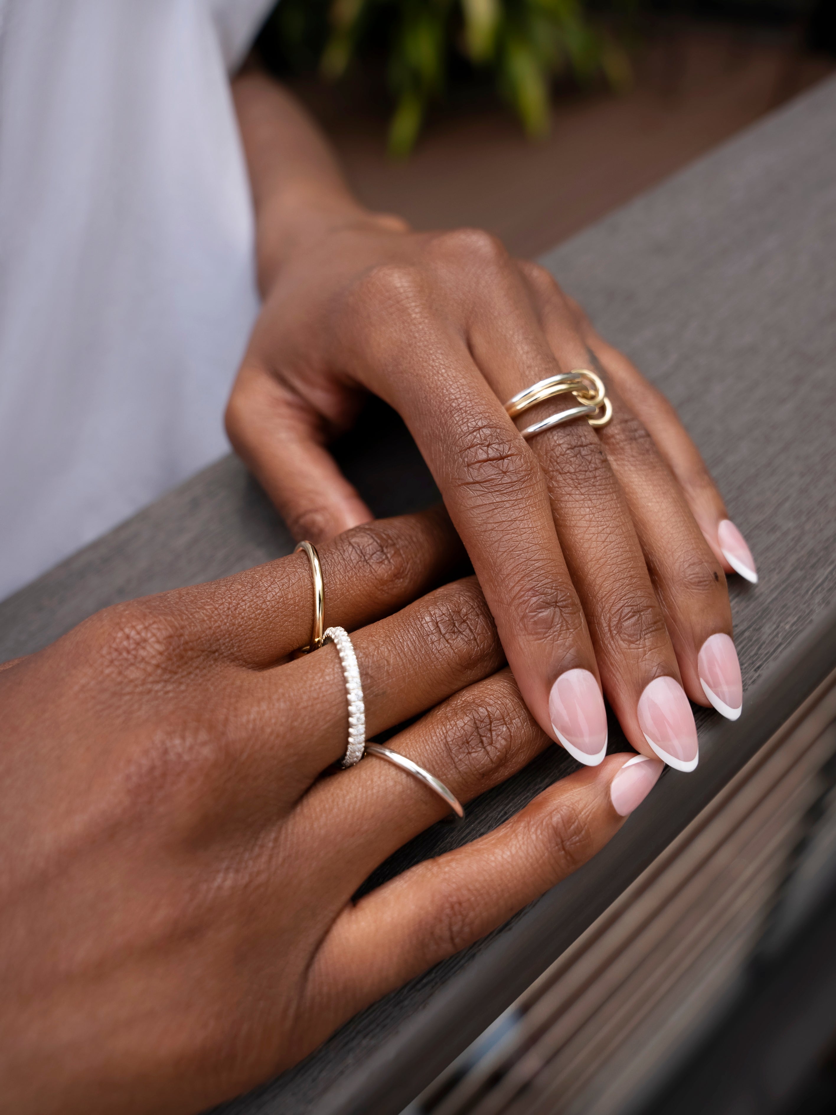 A close-up of hands with a light pink French manicure resting on a railing, adorned with multiple gold and pavé diamond rings, including the stunning Tigris Pavé Band Ring by SPINELLI KILCOLLIN.