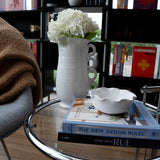 A white vase with white flowers sits on a glass side table beside two books and THE CONRAN SHOP’s Puglia Scalloped Small Bowl; a cozy chair and bookshelves filled with colorful books are in the background.