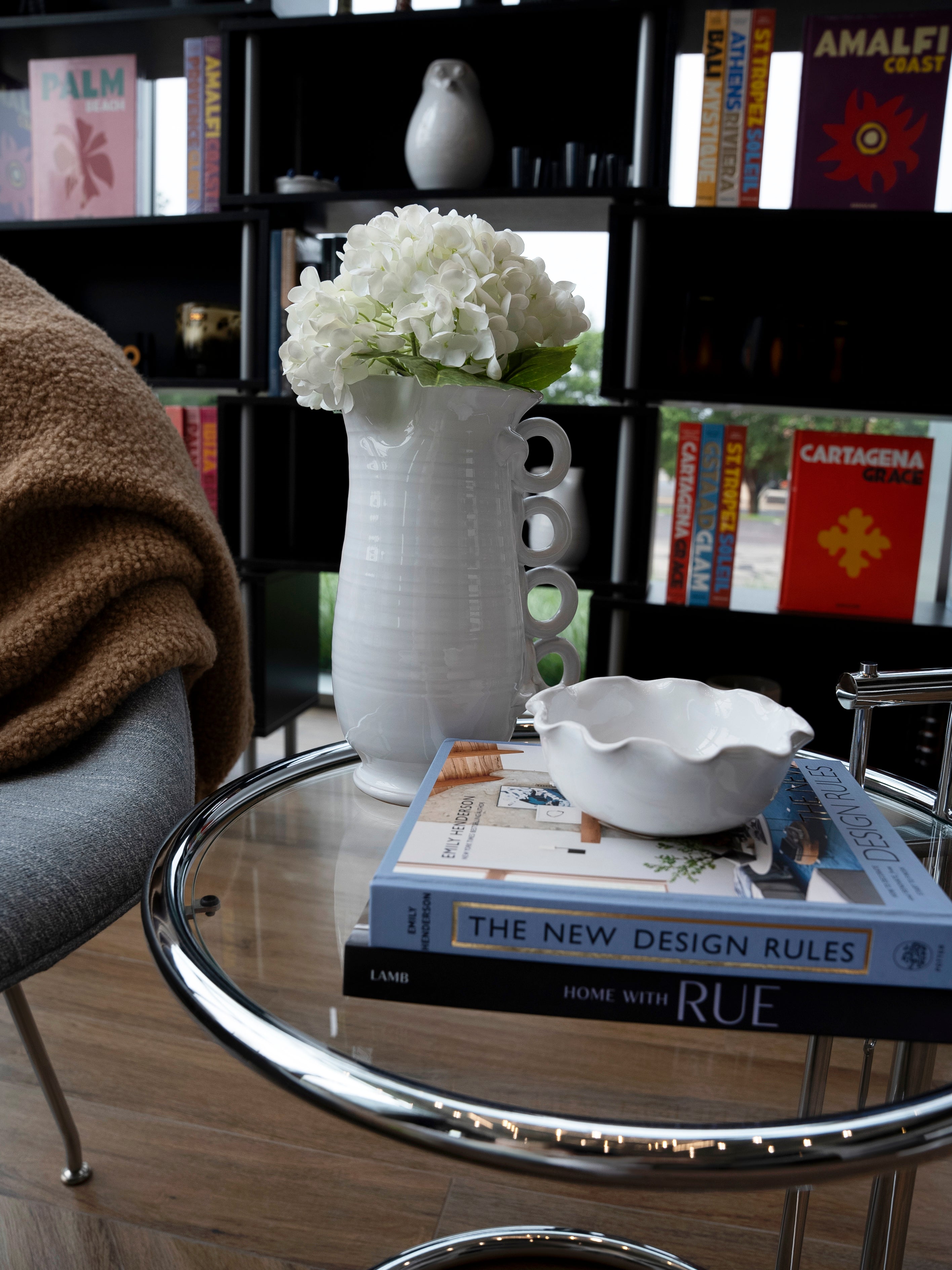 A white vase with white flowers sits on a glass side table beside two books and THE CONRAN SHOP’s Puglia Scalloped Small Bowl; a cozy chair and bookshelves filled with colorful books are in the background.