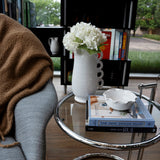 A cozy living room corner features a gray chair draped with a brown blanket, accompanied by a glass side table displaying books and THE CONRAN SHOP’s Puglia Jug with Ring Handle—a handcrafted white stoneware piece—alongside a white vase filled with white flowers and a decorative bowl. In the background, a bookshelf and window view complete the scene.