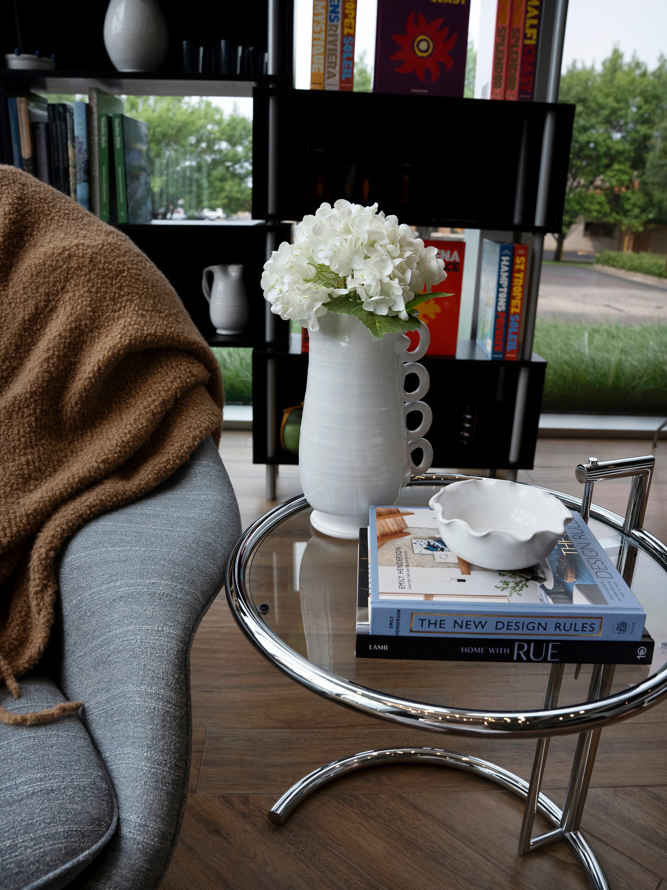 A cozy living room corner features a gray chair draped with a brown blanket, accompanied by a glass side table displaying books and THE CONRAN SHOP’s Puglia Jug with Ring Handle—a handcrafted white stoneware piece—alongside a white vase filled with white flowers and a decorative bowl. In the background, a bookshelf and window view complete the scene.