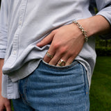 A person wearing a light button-up shirt and blue jeans stands with one hand in their pocket, displaying several rings—including the striking SPINELLI KILCOLLIN Phantom SK Ring—and a chain bracelet.