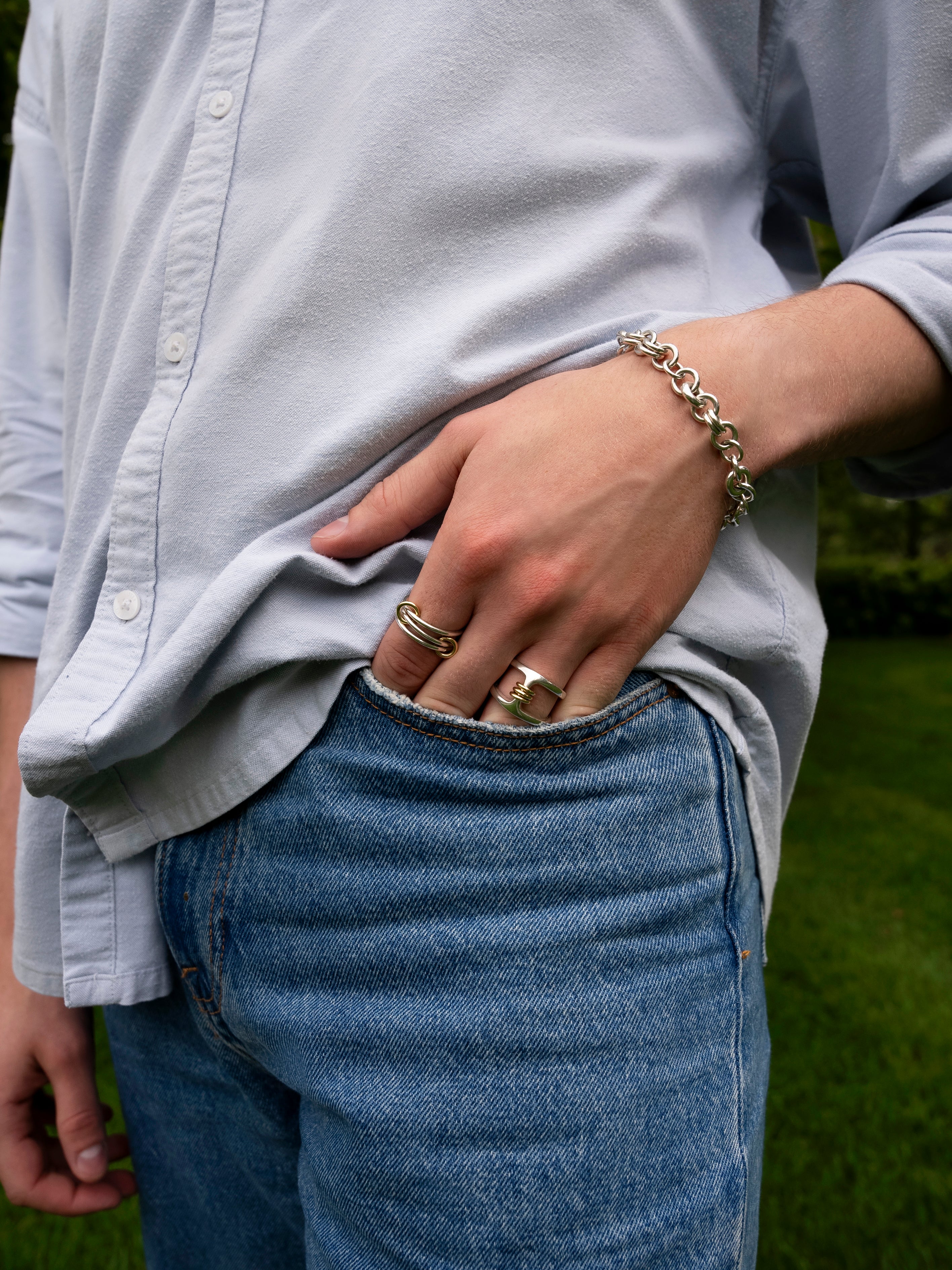 A person wearing a light button-up shirt and blue jeans stands with one hand in their pocket, displaying several rings—including the striking SPINELLI KILCOLLIN Phantom SK Ring—and a chain bracelet.