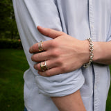 A person in a light blue shirt with rolled-up sleeves stands outdoors, wearing silver rings—including the Phantom SK Ring by SPINELLI KILCOLLIN—and a chain bracelet on their left hand, which rests on their right arm. Grass and trees are visible in the background.