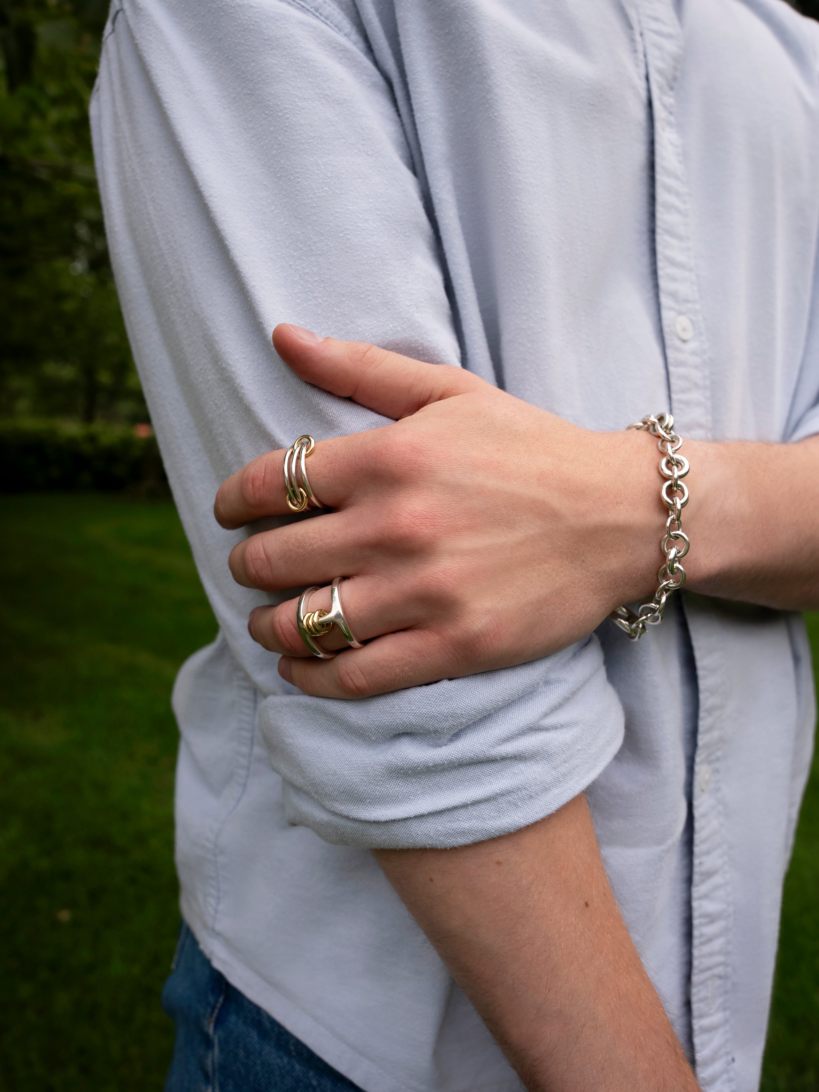 A person in a light blue shirt with rolled-up sleeves stands outdoors, wearing silver rings—including the Phantom SK Ring by SPINELLI KILCOLLIN—and a chain bracelet on their left hand, which rests on their right arm. Grass and trees are visible in the background.